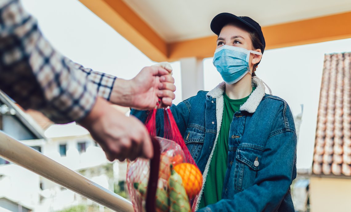 A young adult wearing a mask is handing a bag of groceries to an older adult.