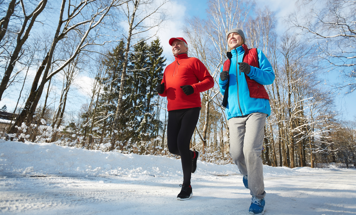 Happy active seniors in sportswear jogging down winter road in natural environment