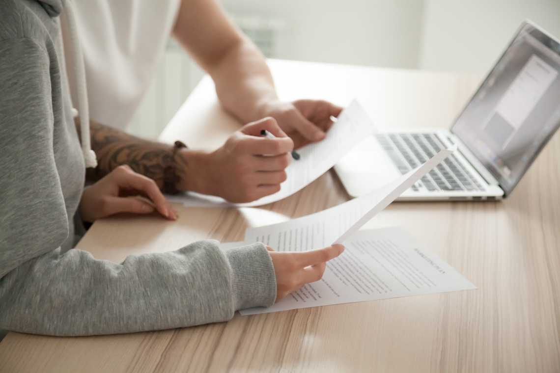 Close up of two people at a desk reviewing important documents. Only their arms and torsos are visible and there are papers and a laptop in front of them.