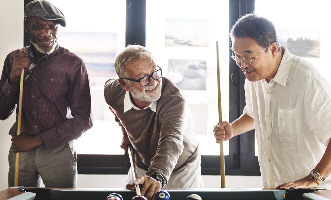 Three retirees are relaxed and enjoying playing a game of pool together. One person is lining up their shot as the other two look on.