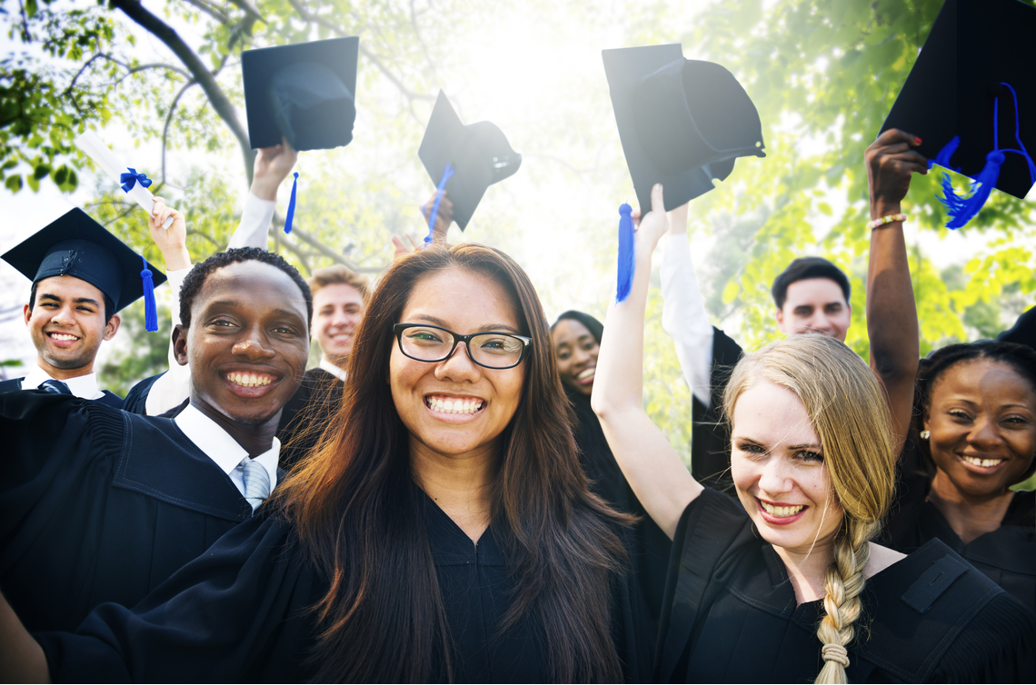 A diverse group of friends celebrating outside on a sunny day after their university commencement. They're wearing graduation gowns, smiling broadly and some are holding their grad caps above their heads.