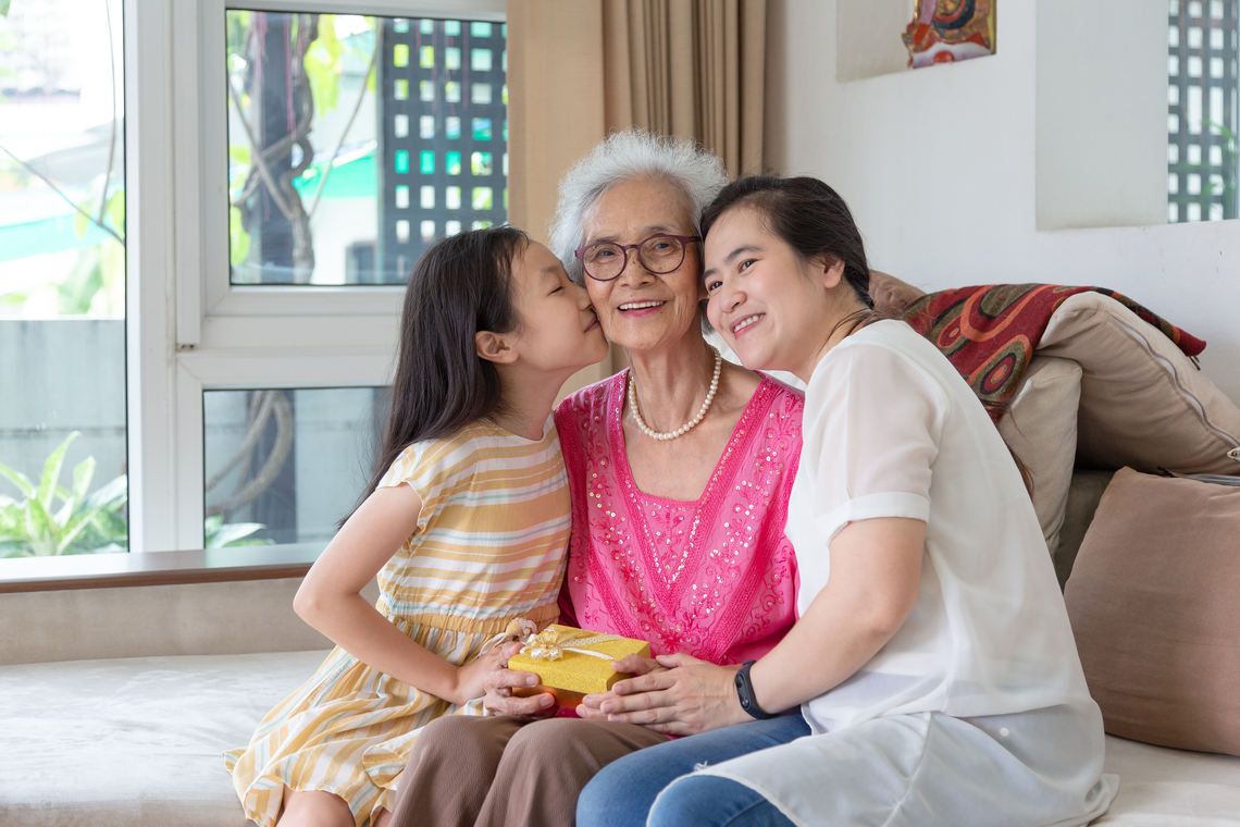Trois générations d’une même famille posent pour une photo – la grand-mère, sa fille et sa petite-fille. La grand-mère au centre regarde la caméra et sa petite-fille à gauche l’embrasse sur la joue. 
