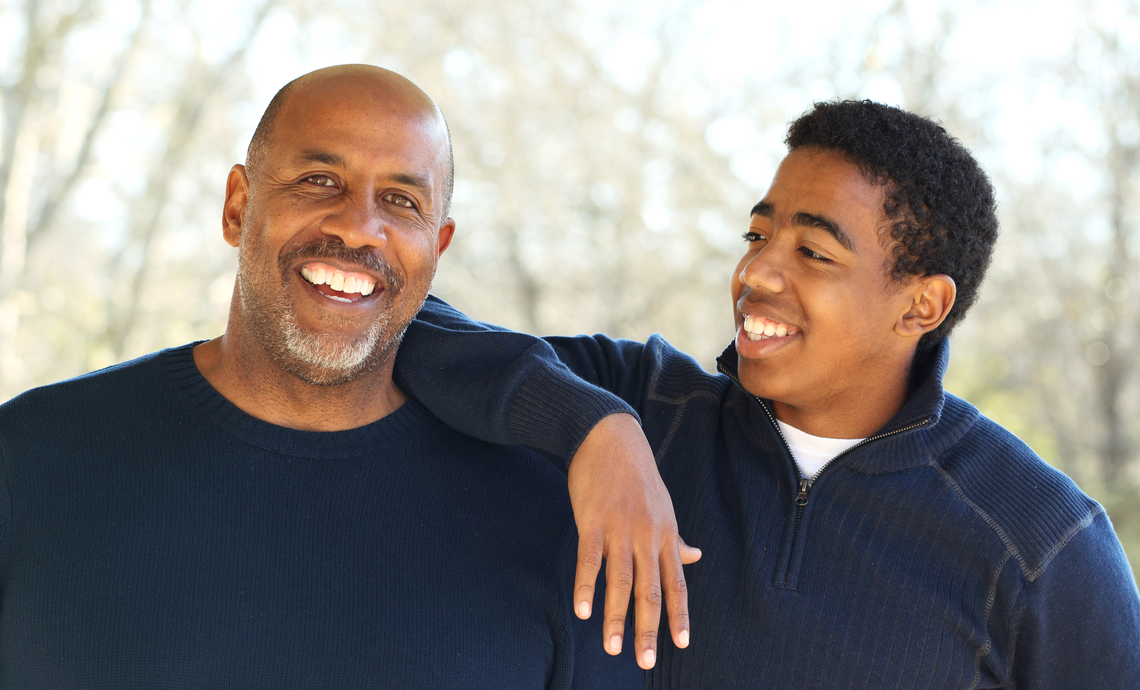 A father and teenage son. The son is smiling at his dad with his arm resting on his dad’s shoulder