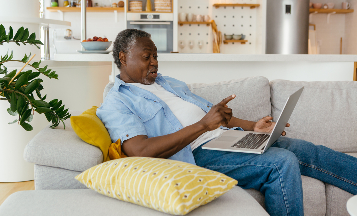 Older adult sitting on a couch and chatting with a friend over video with a laptop
