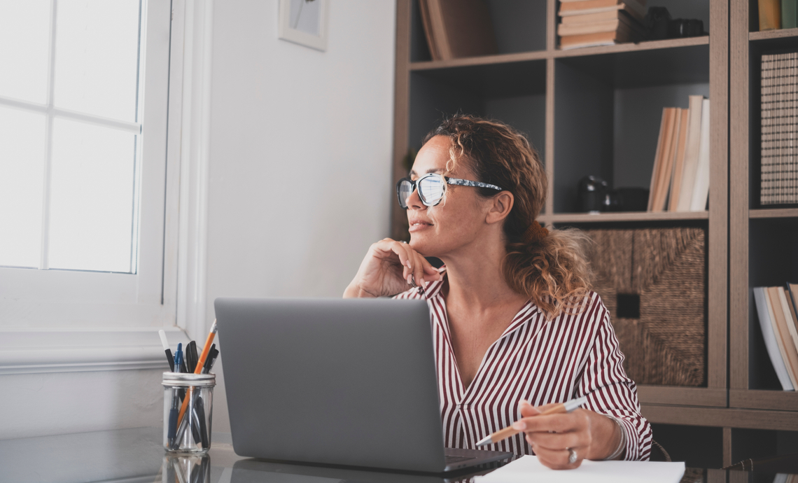 Teacher sitting in front of a laptop at a desk and holding a pen above a paper pad staring out a window thinking