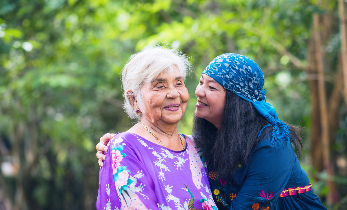 Older parent and adult child sitting outside, the adult child has her arm around the parent and is looking at her lovingly.
