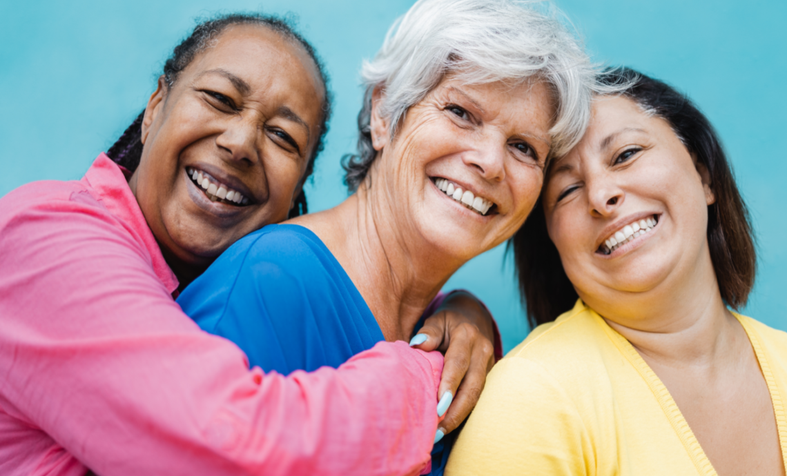 Photo de trois femmes âgées qui sourient. La femme à gauche étreint par derrière celle du centre.