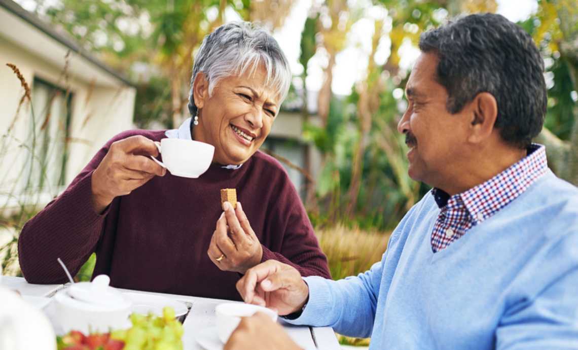 De vieux amis assis à une table dehors parlent et rient tout en dégustant du thé et des fruits.