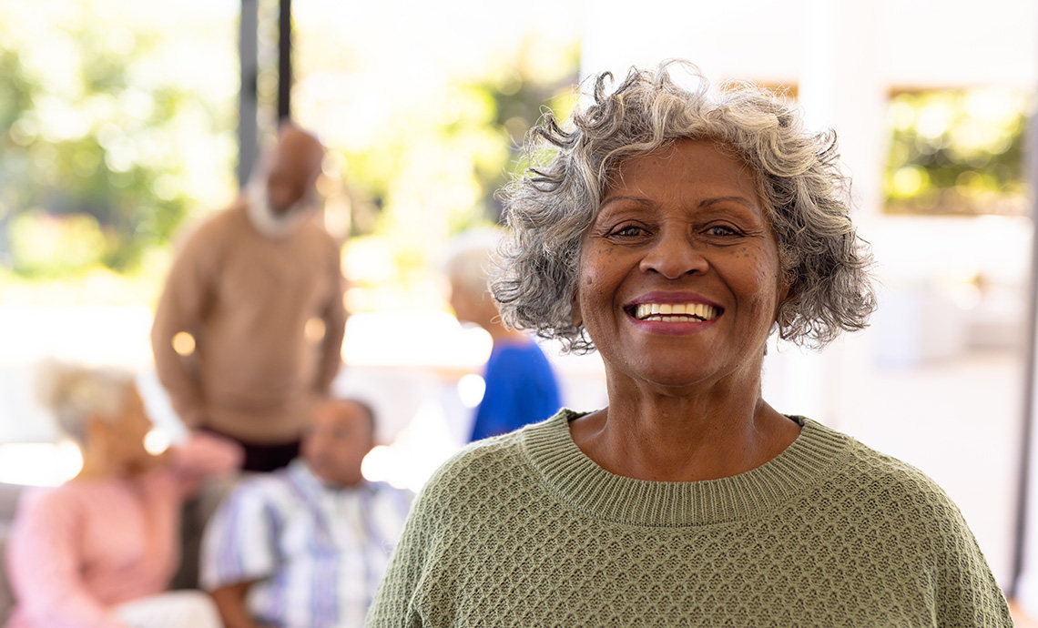 Smiling older woman with grey curly hair and a green sweater. A group of friends are hanging out in the background. 