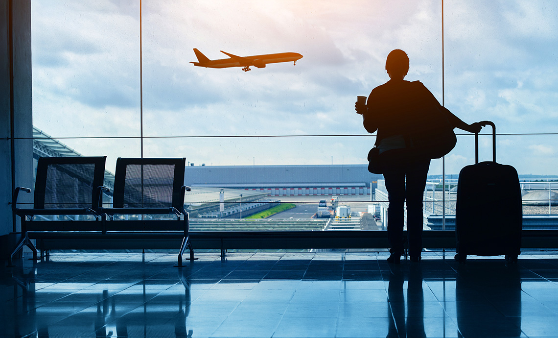 Silhouette of a person at an airport window holding a coffee and suitcase, watching a plane take off.