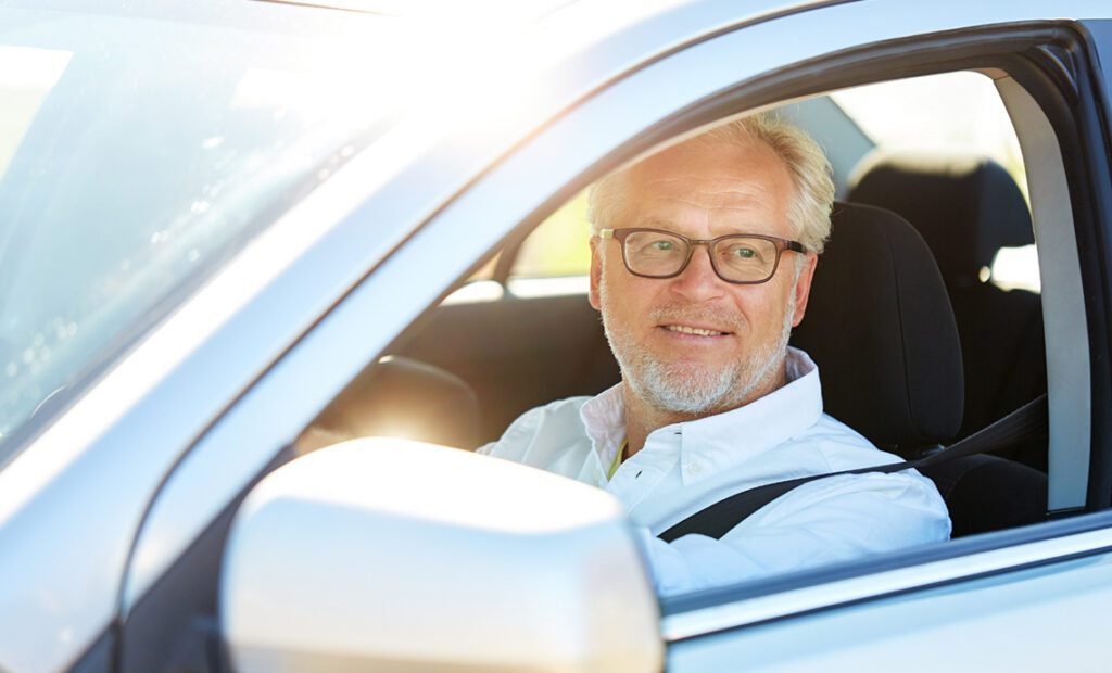 Homme âgé portant des verres et une chemise blanche à col ouvert, conduisant une voiture grise.